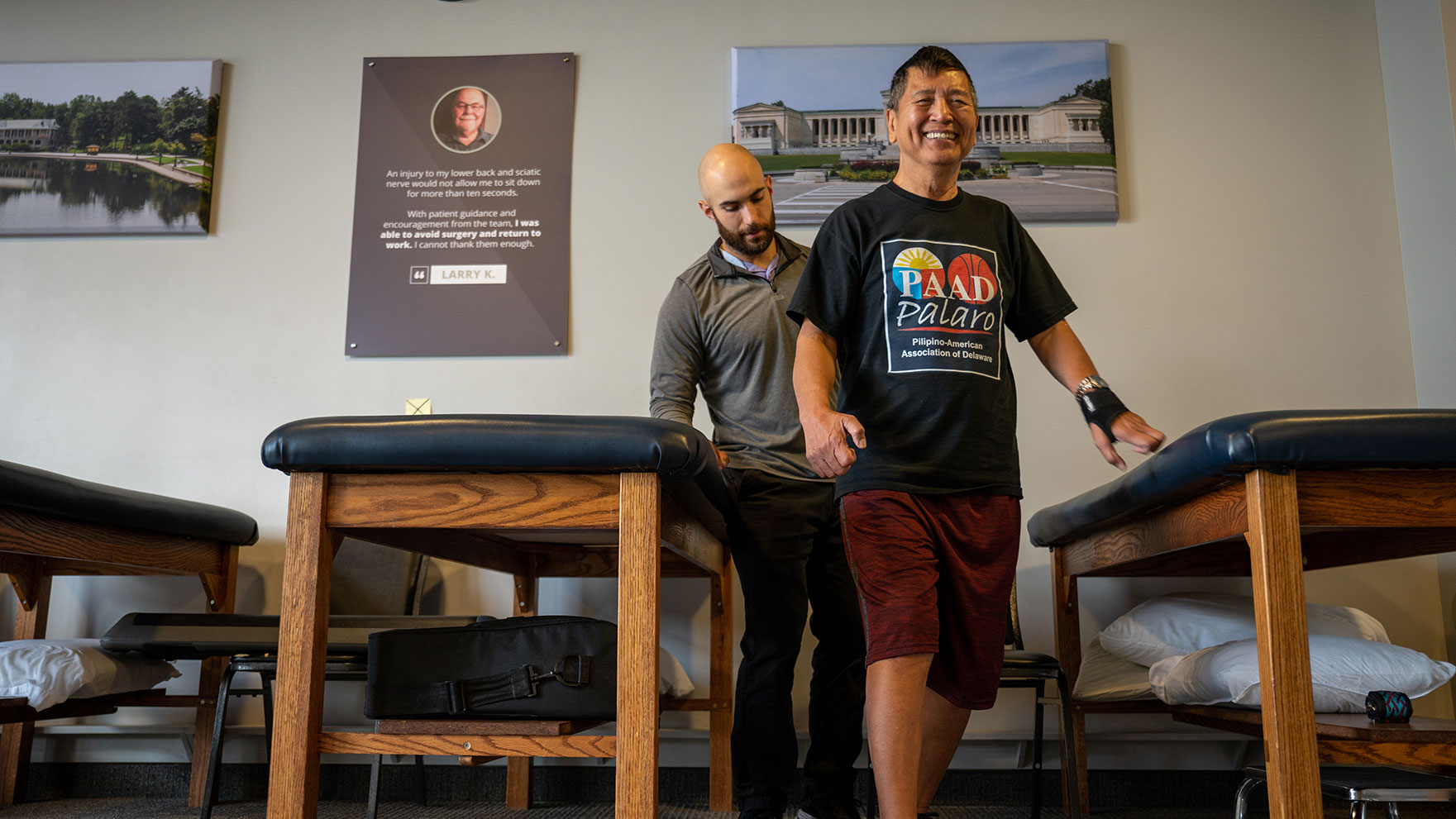 A physical therapist smiles as his patient walks before him.