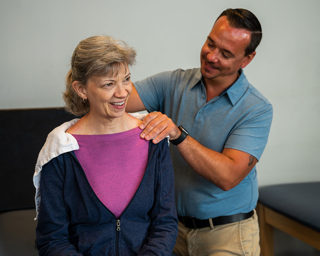 A physical therapist assistant smiles as he massages the shoulder of his patient.