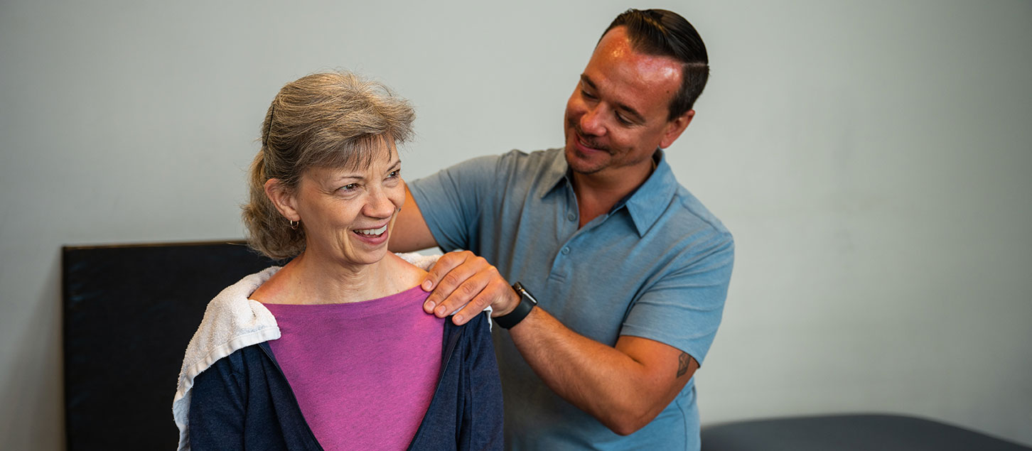 A physical therapist assistant smiles as he massages the shoulder of his patient.