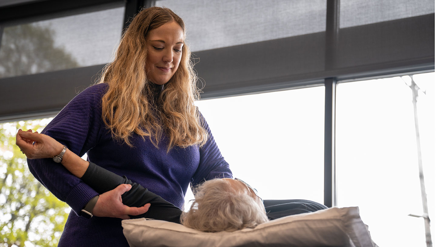 A blond physical therapist smiles as she stretches the arm of her patient.