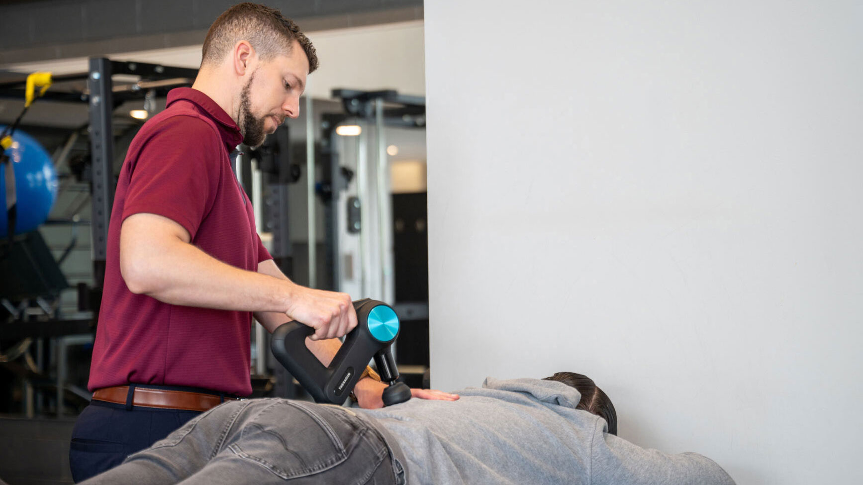A Physical Therapist uses a massage gun to treat his patient who lies on a table