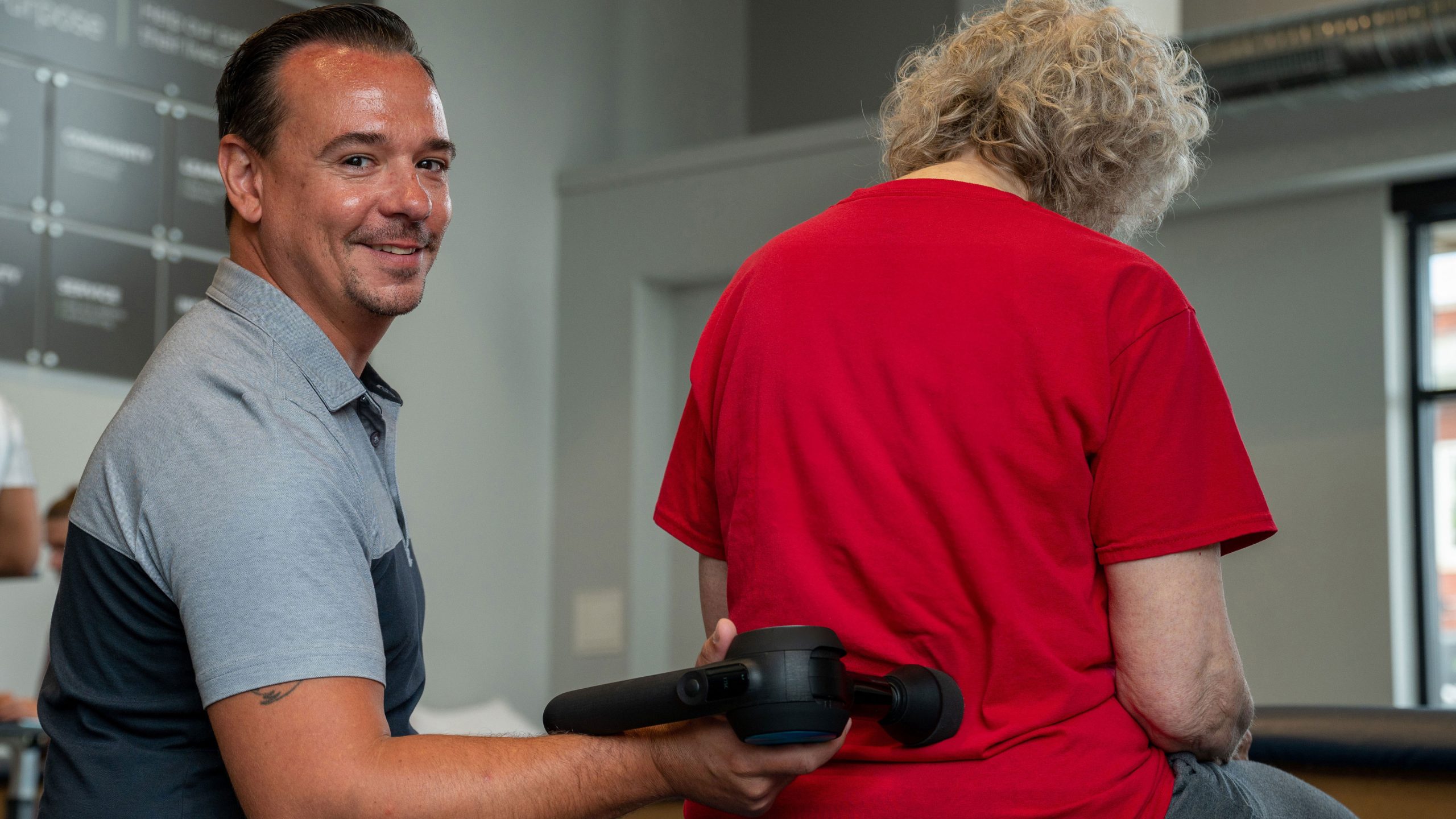 A Physical Therapist smiles at the camera as he uses a massage gun on his patient.