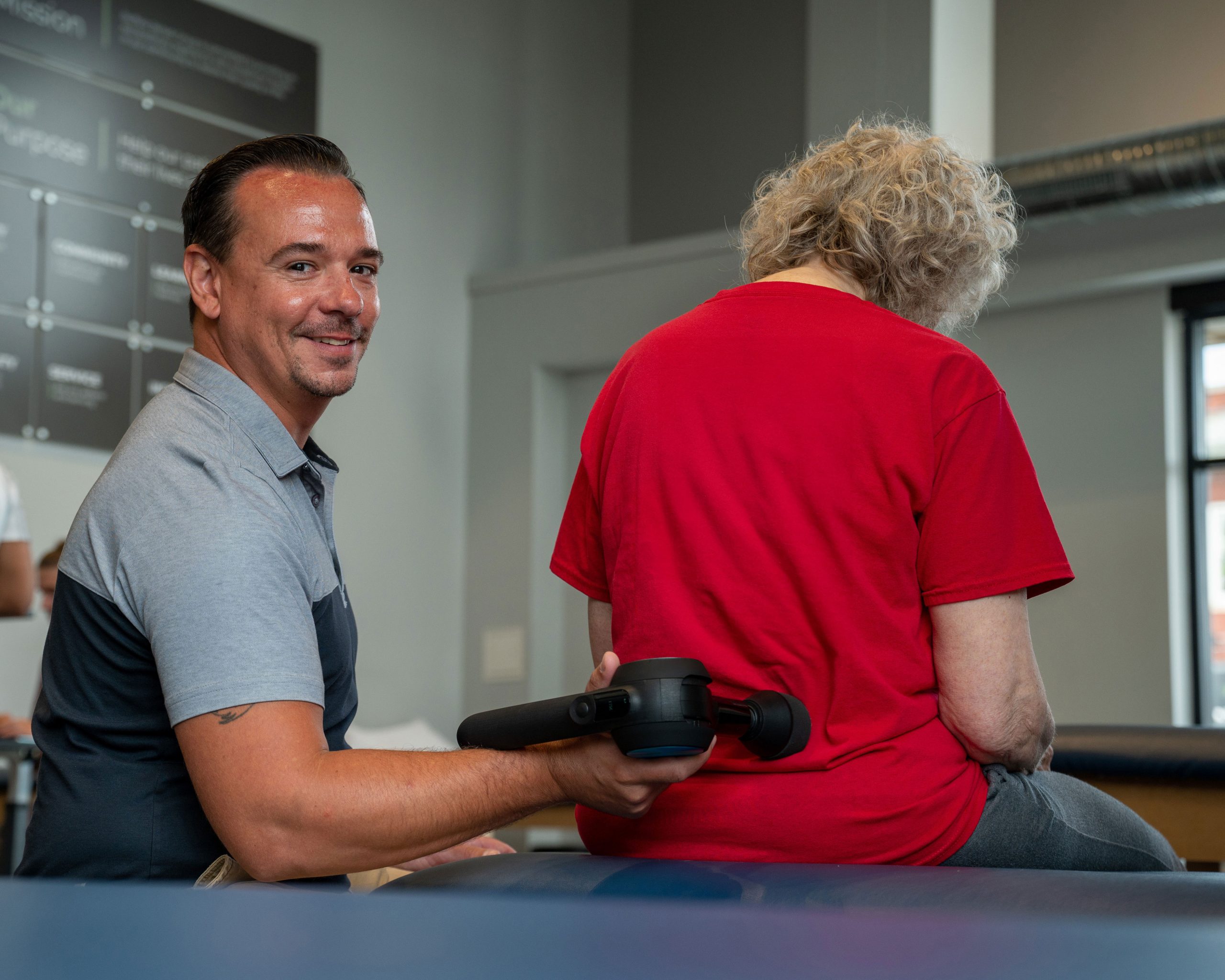 A Physical Therapist smiles at the camera as he uses a massage gun on his patient.