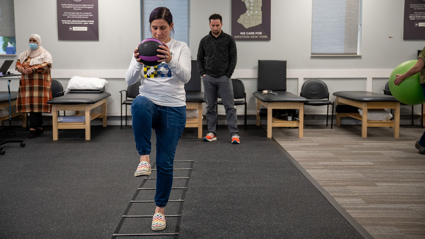 A physical therapist watches as his patient walks with a medicine ball.
