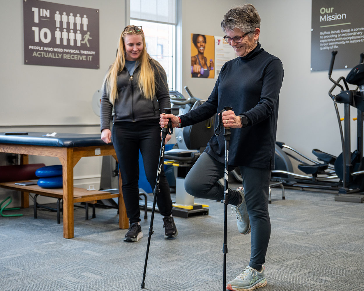 A physical therapost smiles as she watches her patient walk with the aid of crutches.