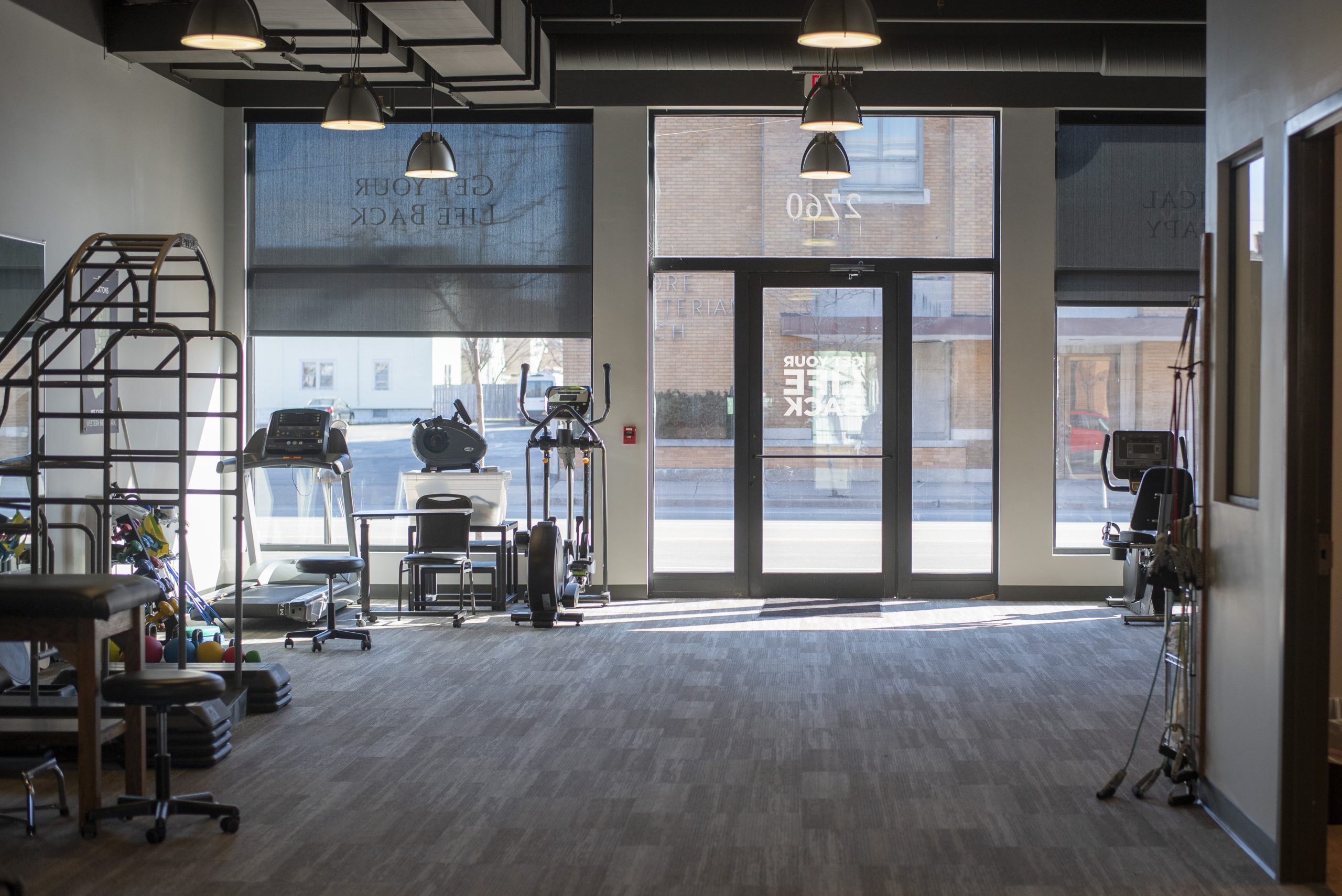 A Physical Therapy clinic with the sun coming in onto gray carpet. There are white walls and various pieces of therapy equipment.