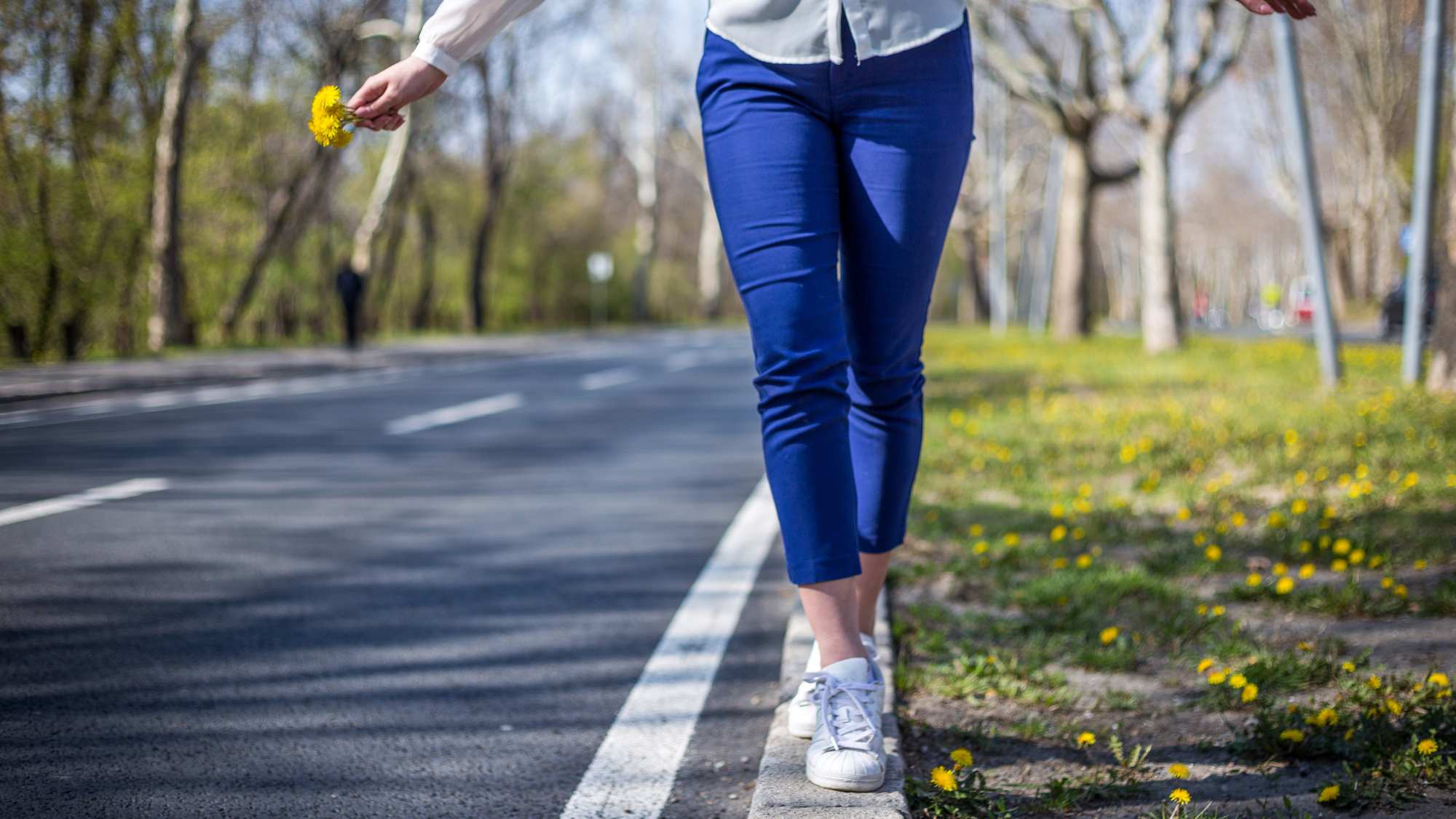 An image of a woman from the waist down walking and balancing on the white line at the side of a road. She has yellow flowers in her hand.