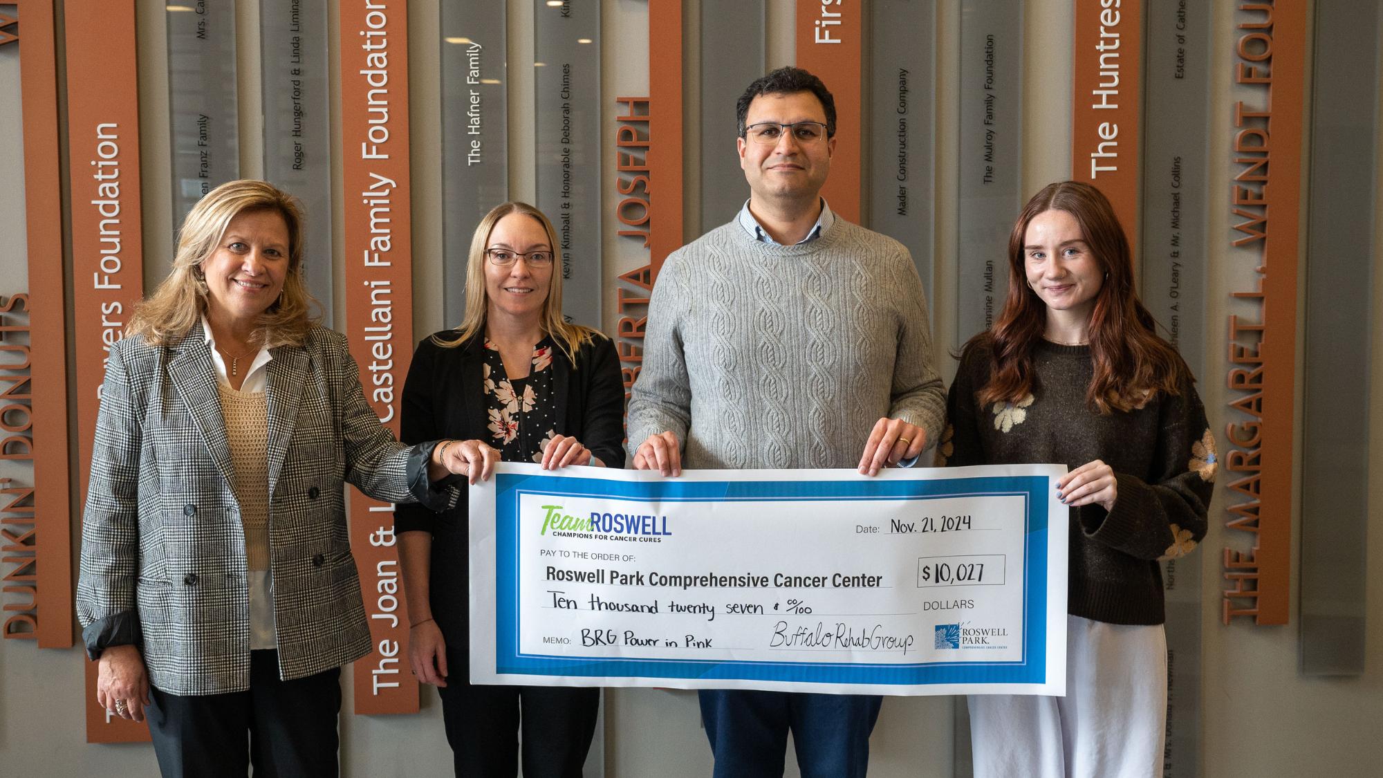 An image of four people, three female and one male, holding a large check valued at $10,027. It's a ceremonial check, signifying a donation.